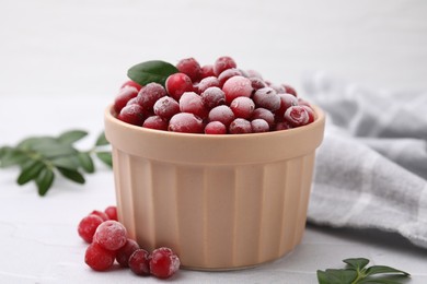 Photo of Frozen red cranberries in bowl and green leaves on white table, closeup