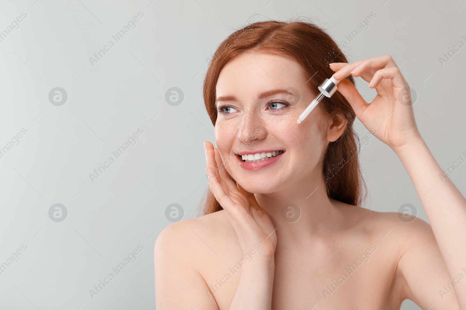 Photo of Smiling woman with freckles applying cosmetic serum onto her face against grey background. Space for text