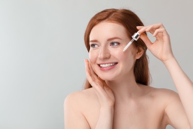 Smiling woman with freckles applying cosmetic serum onto her face against grey background. Space for text