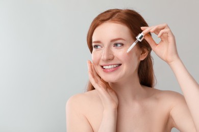 Photo of Smiling woman with freckles applying cosmetic serum onto her face against grey background. Space for text