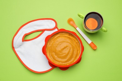 Photo of Baby bib and plastic dishware with healthy food on light green background, flat lay