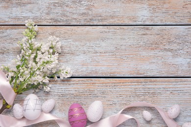 Many painted Easter eggs, branch of lilac flowers and ribbon on rustic wooden table, flat lay. Space for text