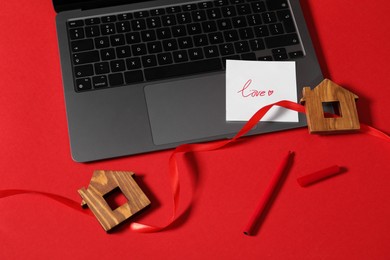 Photo of Flat lay composition with love note and laptop on red background. Ribbon between two wooden house models symbolizing connection in long-distance relationship