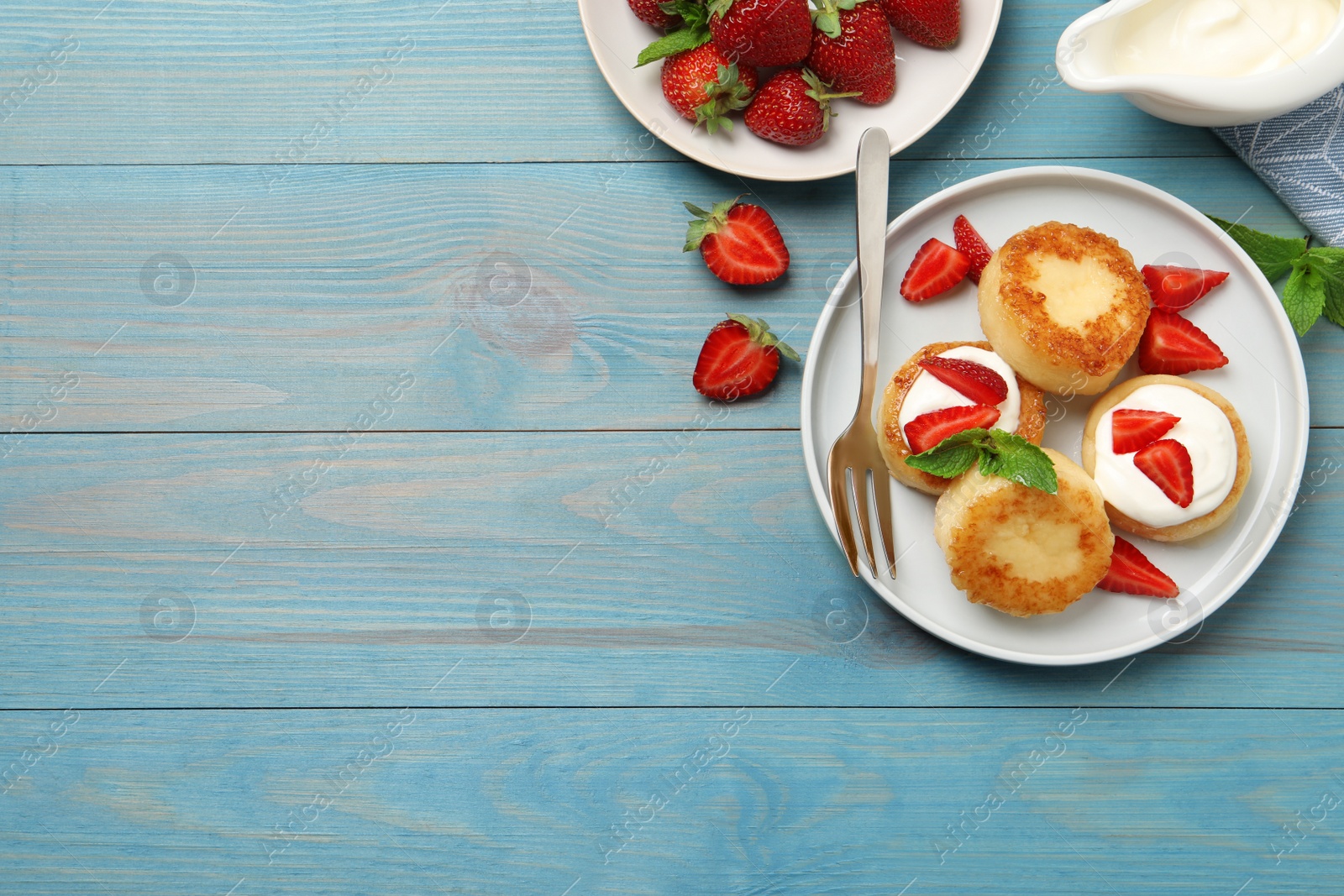 Photo of Delicious cottage cheese pancakes with strawberries, honey and sour cream on light blue wooden table, flat lay. Space for text