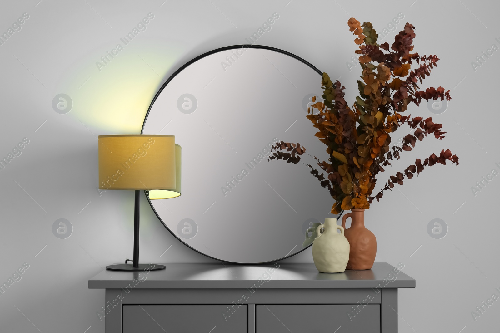 Photo of Stylish round mirror, vases, dried eucalyptus branches and table lamp on chest of drawers near white wall indoors. Interior design