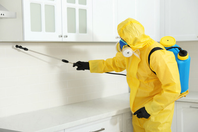 Photo of Pest control worker in protective suit spraying insecticide on furniture indoors