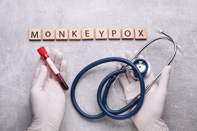 Photo of Word Monkeypox made of wooden squares, top view. Doctor holding test tube and stethoscope at light grey table, closeup