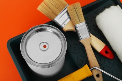 Photo of Can of orange paint, brushes, roller and container on color background, closeup