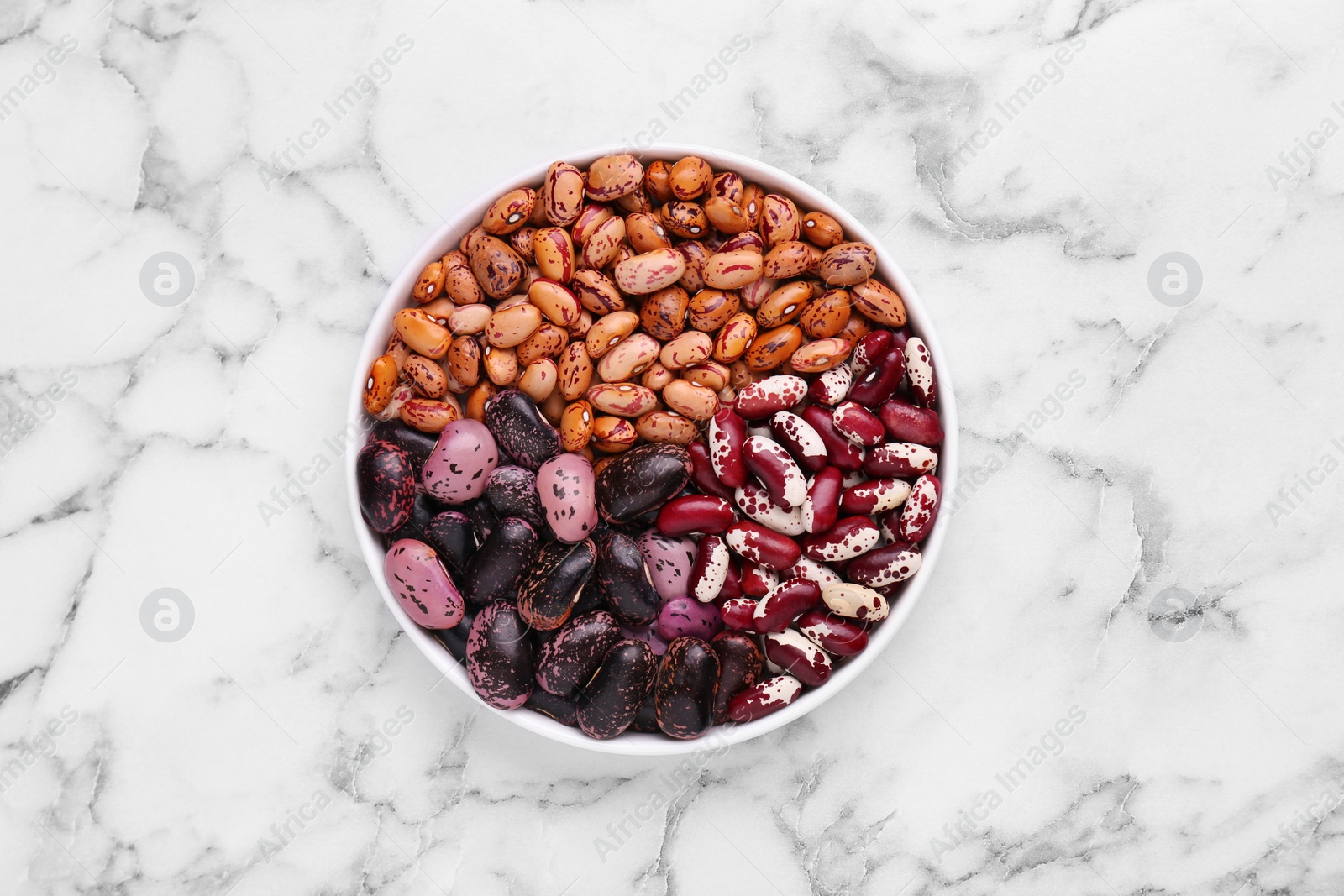 Photo of Different kinds of dry kidney beans in bowl on white marble table, top view