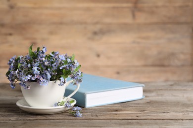 Photo of Beautiful forget-me-not flowers in cup, saucer and book on wooden table, closeup. Space for text