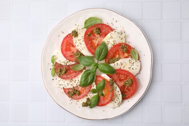 Plate of delicious Caprese salad with herbs on white tiled table, top view