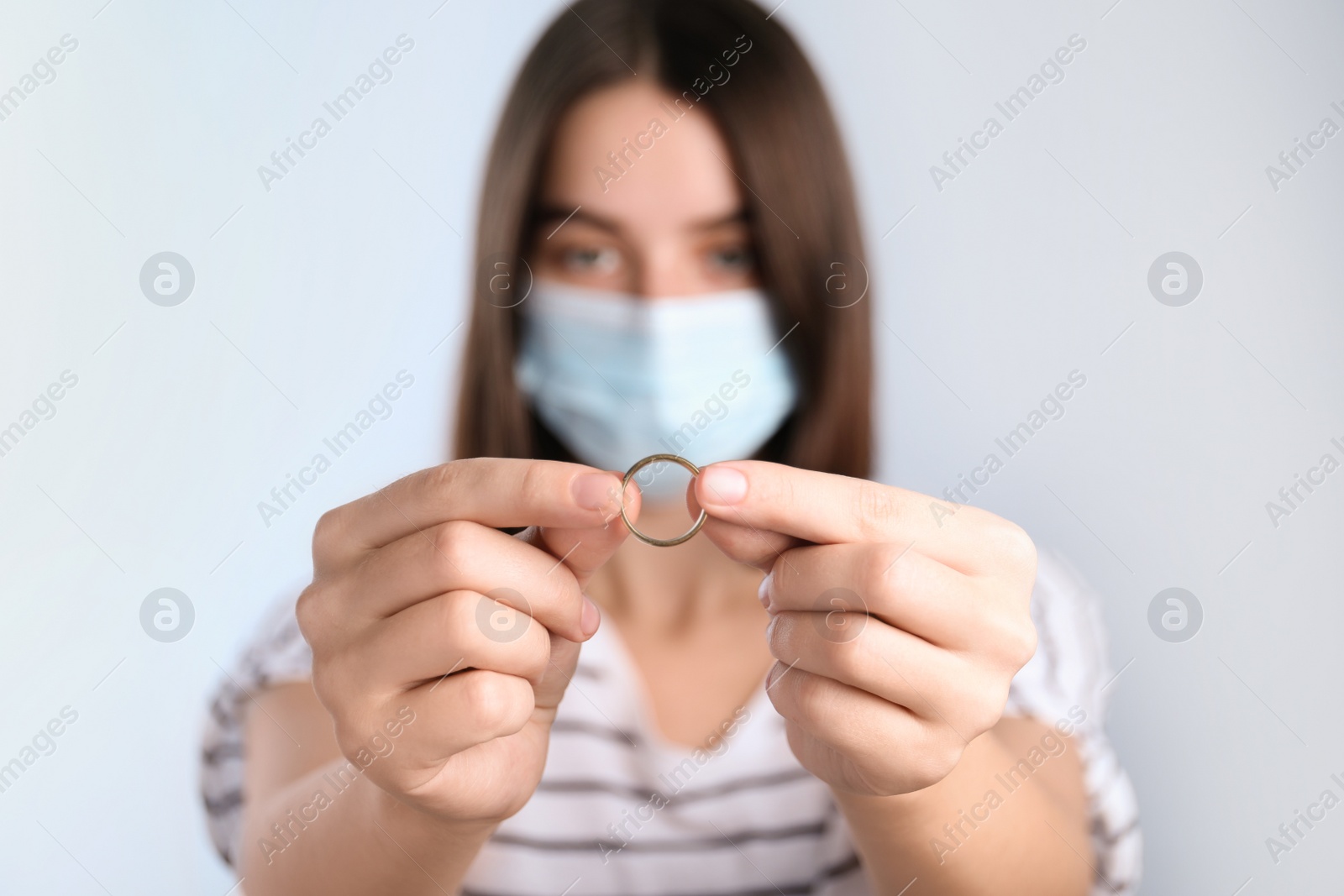 Photo of Woman in protective mask holding wedding ring against light background, focus on hands. Divorce during coronavirus quarantine