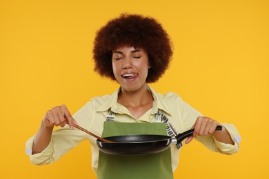 Photo of Happy young woman in apron holding frying pan and spatula on orange background