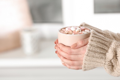 Photo of Woman holding cup of aromatic cacao with marshmallows on blurred background, closeup. Space for text