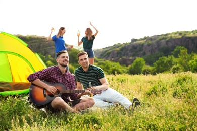 Photo of Group of young people resting with beer and guitar near camping tent in wilderness