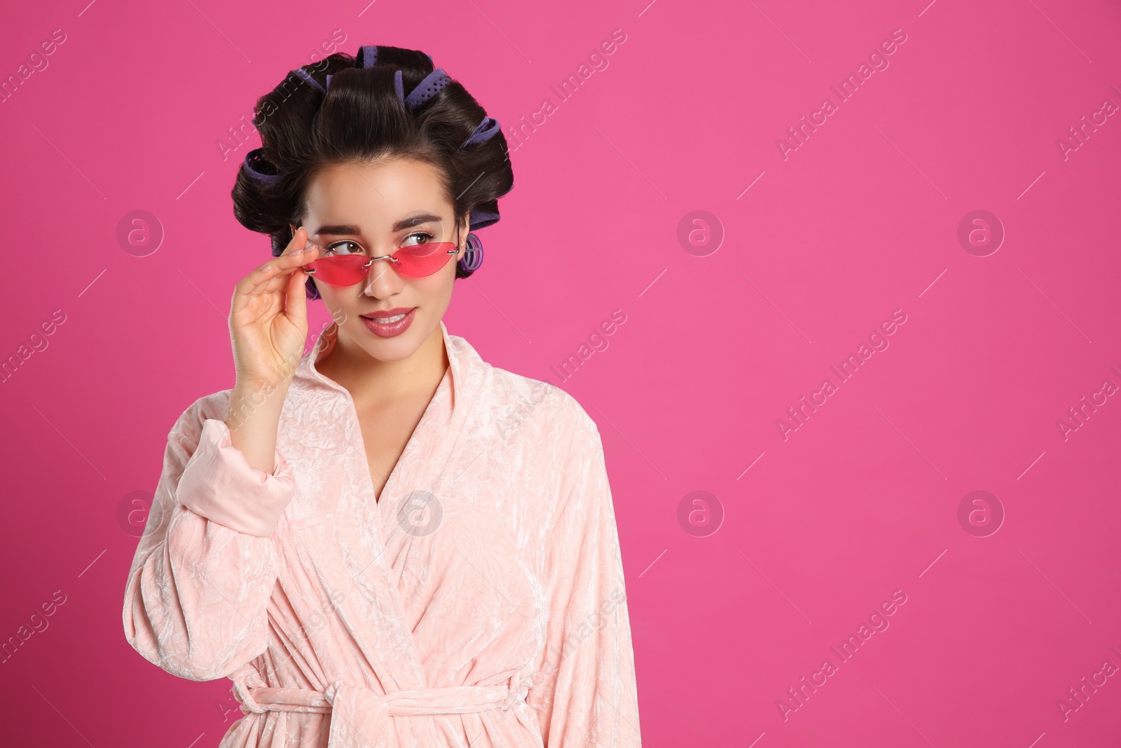 Photo of Young woman in bathrobe with hair curlers and sunglasses on pink background, space for text