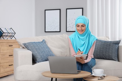 Photo of Muslim woman writing notes near laptop at couch in room. Space for text