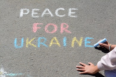 Photo of Girl writing Peace For Ukraine with colorful chalks on asphalt outdoors, closeup