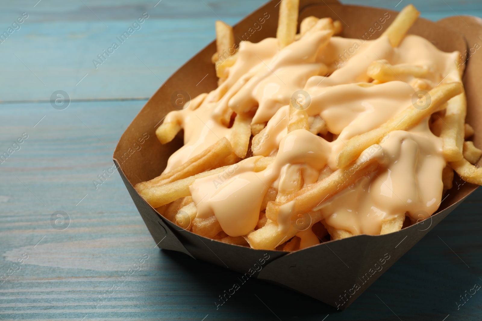 Photo of Tasty potato fries and cheese sauce in paper container on light blue wooden table, closeup