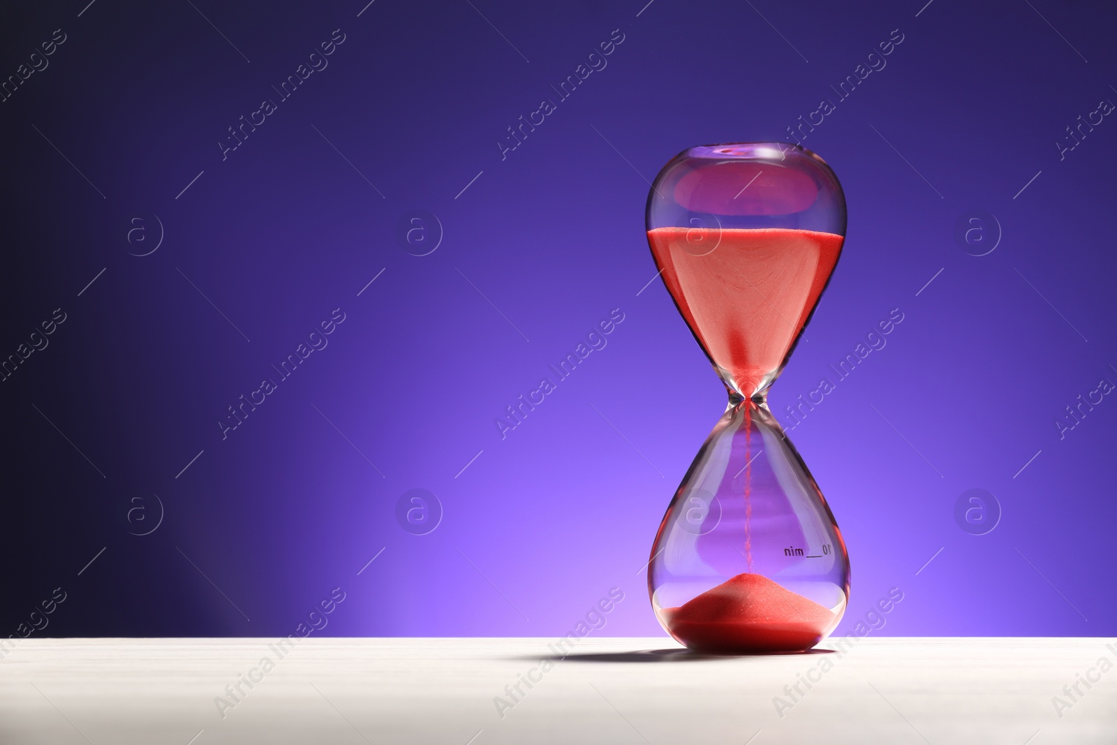 Photo of Hourglass with red flowing sand on white table against purple background. Space for text