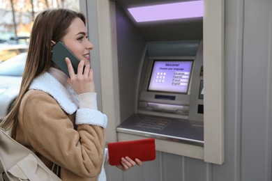 Image of Young woman talking by mobile phone while using cash machine for money withdrawal outdoors