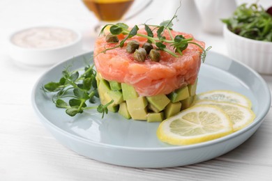 Photo of Tasty salmon tartare with avocado, lemon, capers and microgreens on white wooden table, closeup