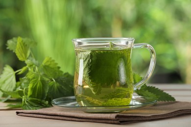 Photo of Glass cup of aromatic nettle tea and green leaves on wooden table outdoors