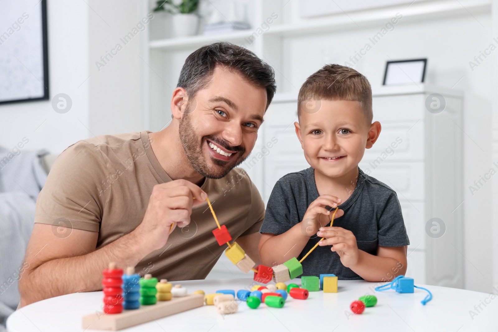 Photo of Motor skills development. Father and his son playing with wooden pieces and string for threading activity at table indoors