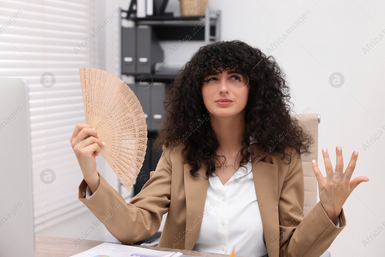 Photo of Young businesswoman waving hand fan to cool herself at table in office