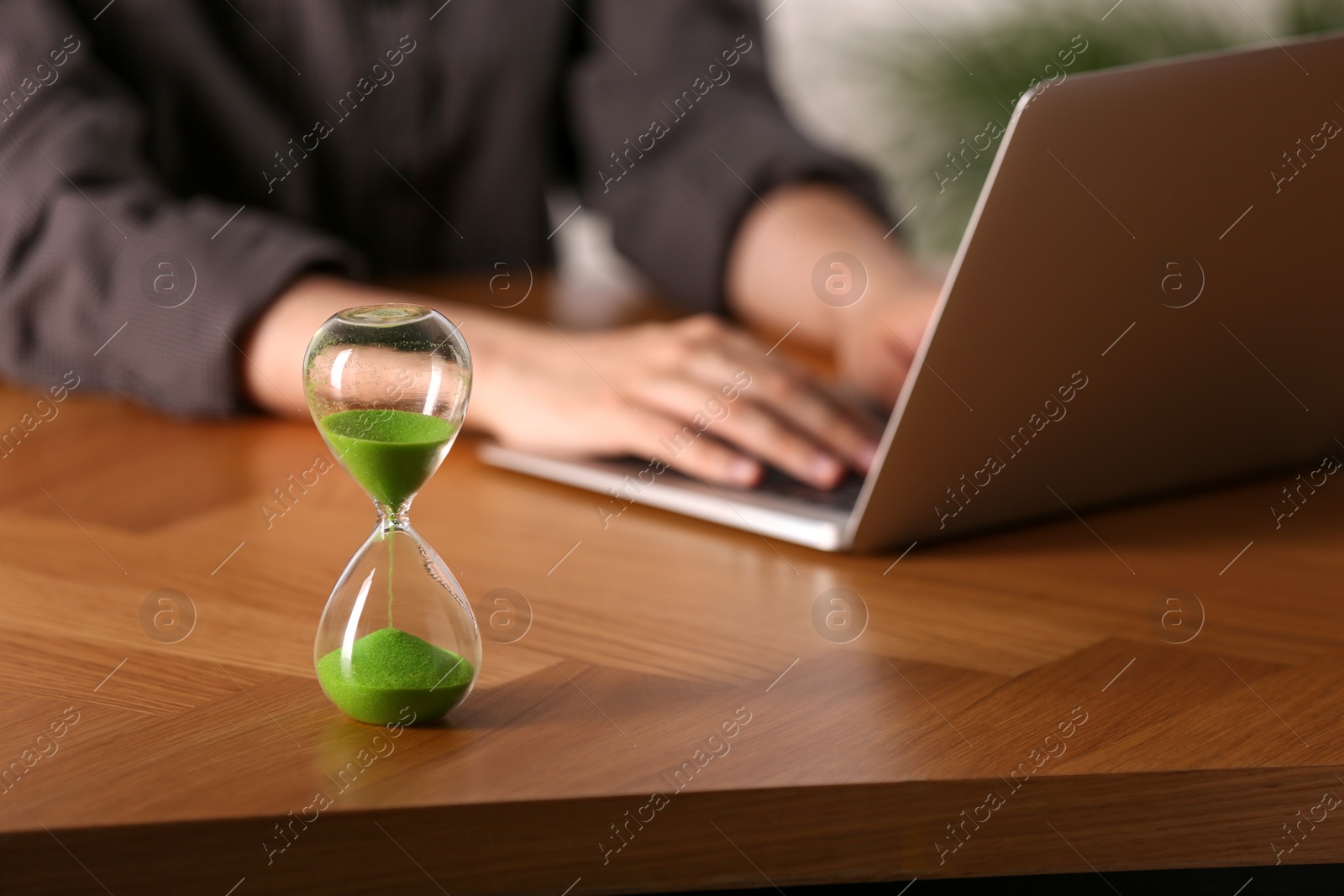 Photo of Hourglass with flowing sand on wooden table, selective focus. Man using laptop indoors