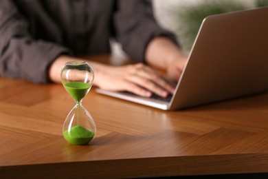 Photo of Hourglass with flowing sand on wooden table, selective focus. Man using laptop indoors