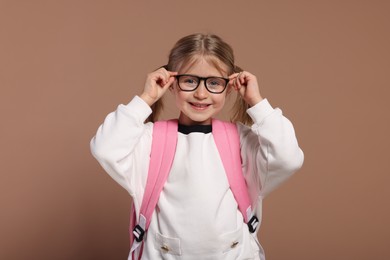 Happy schoolgirl in glasses with backpack on brown background