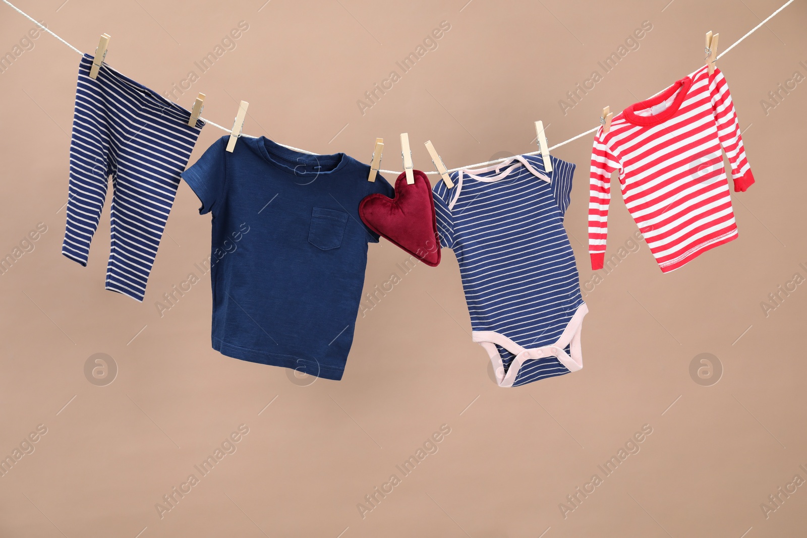 Photo of Different baby clothes and heart shaped cushion drying on laundry line against light brown background