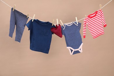 Different baby clothes and heart shaped cushion drying on laundry line against light brown background