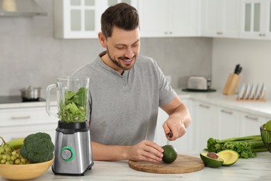 Photo of Happy man cutting avocado for delicious smoothie at white marble table in kitchen