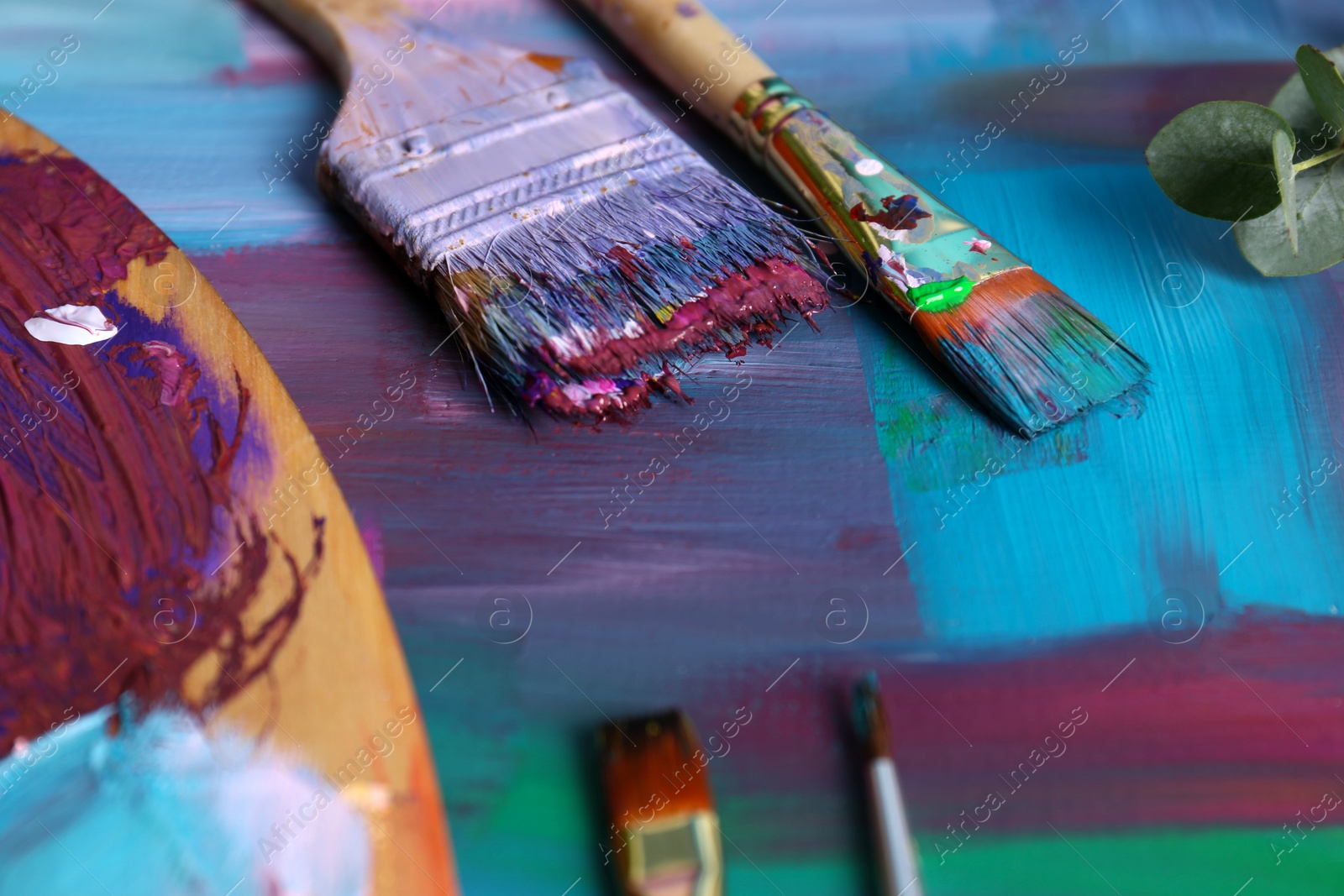 Photo of Canvas with colorful abstract painting, different brushes and wooden artist's palette, closeup