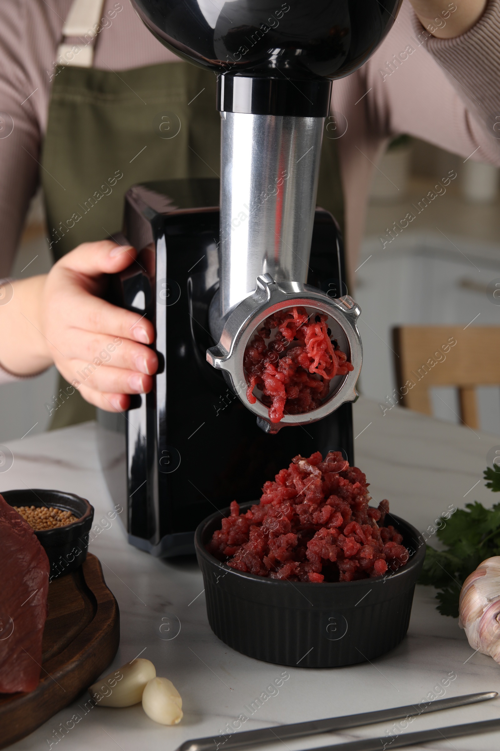 Photo of Woman making beef mince with electric meat grinder at white marble table in kitchen, closeup