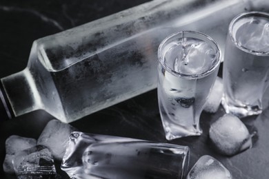 Photo of Bottle of vodka and shot glasses with ice on black table, closeup