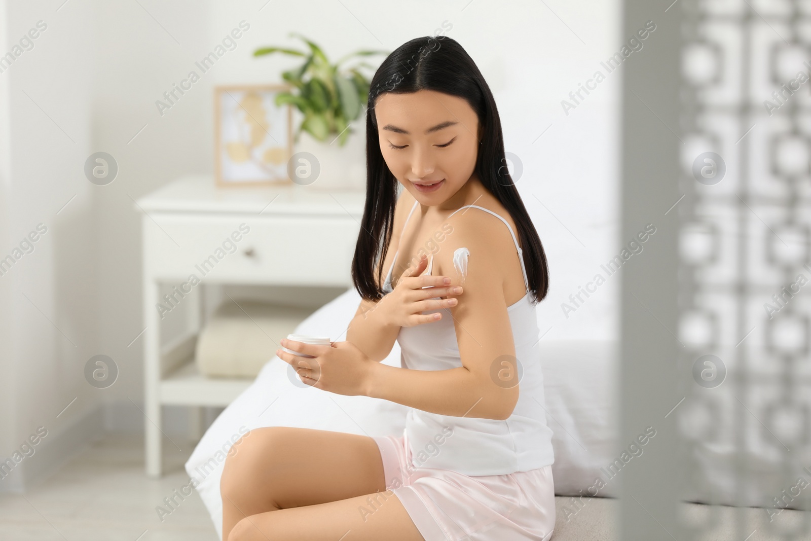 Photo of Beautiful young Asian woman applying body cream on shoulder in bedroom