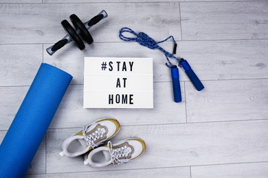 Sport equipment and lightbox with hashtag STAY AT HOME on floor, flat lay. Message to promote self-isolation during COVID‑19 pandemic