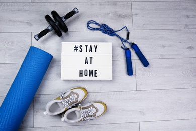 Photo of Sport equipment and lightbox with hashtag STAY AT HOME on floor, flat lay. Message to promote self-isolation during COVID‑19 pandemic