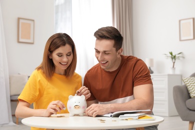 Photo of Couple putting coin into piggy bank at table in living room. Saving money