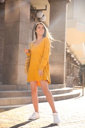 Beautiful young woman in stylish yellow dress with handbag on city street
