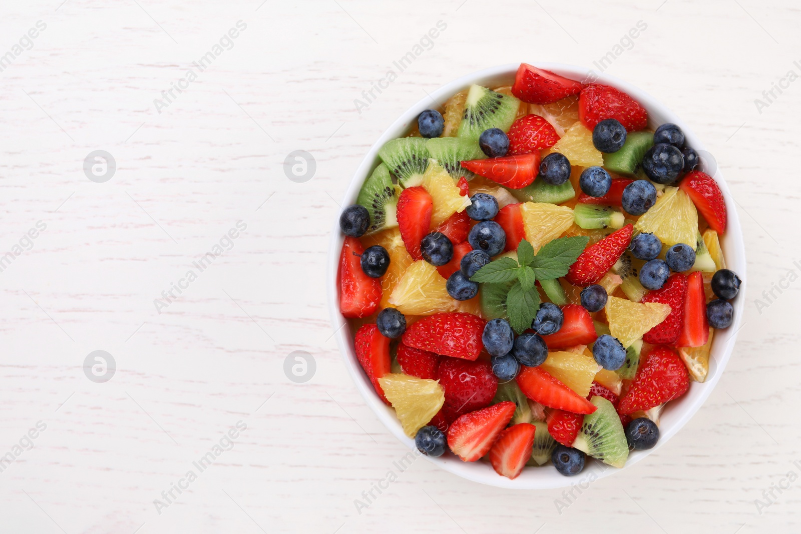 Photo of Delicious fresh fruit salad in bowl on white wooden table, top view. Space for text