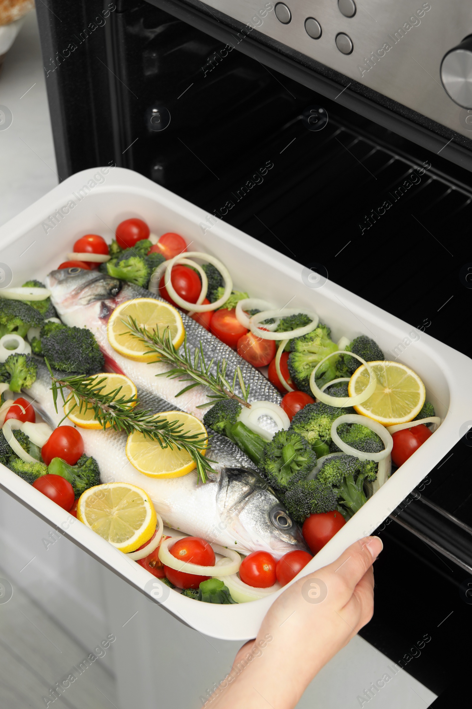 Photo of Woman putting baking dish with raw fish and vegetables into oven, closeup