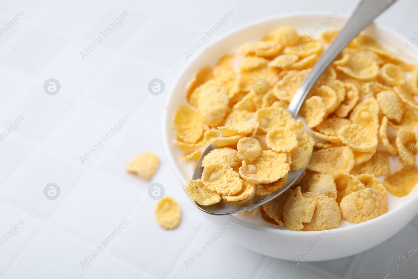 Photo of Breakfast cereal. Tasty corn flakes with milk in bowl and spoon on white tiled table, closeup. Space for text