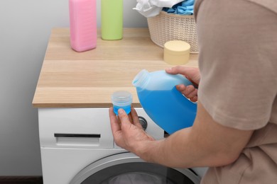 Man pouring fabric softener from bottle into cap near washing machine indoors, closeup