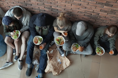 Poor people with plates of food sitting at wall indoors, view from above