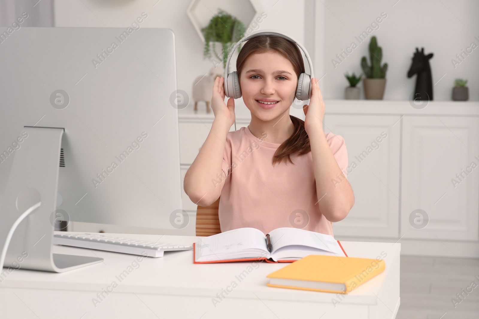 Photo of Cute girl using computer and headphones at desk in room. Home workplace