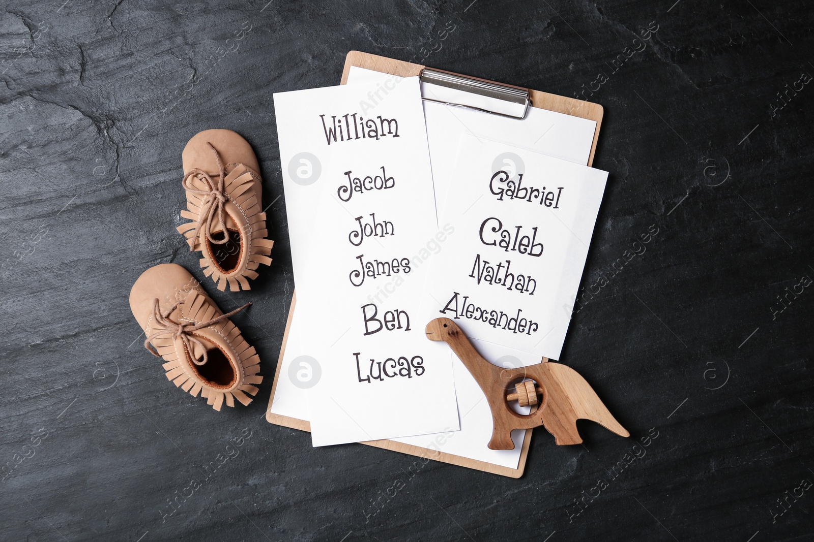 Photo of Clipboard with list of baby names, child's shoes and wooden toy on black table, flat lay